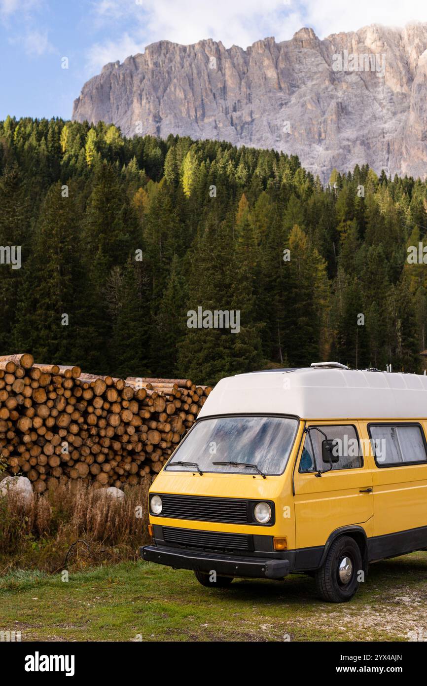 Vintage-Wohnmobil parkt vor gehäuften Holzstapeln mit Berggipfeln und Kiefernwäldern im Hintergrund. Vertikale Ausrichtung Stockfoto