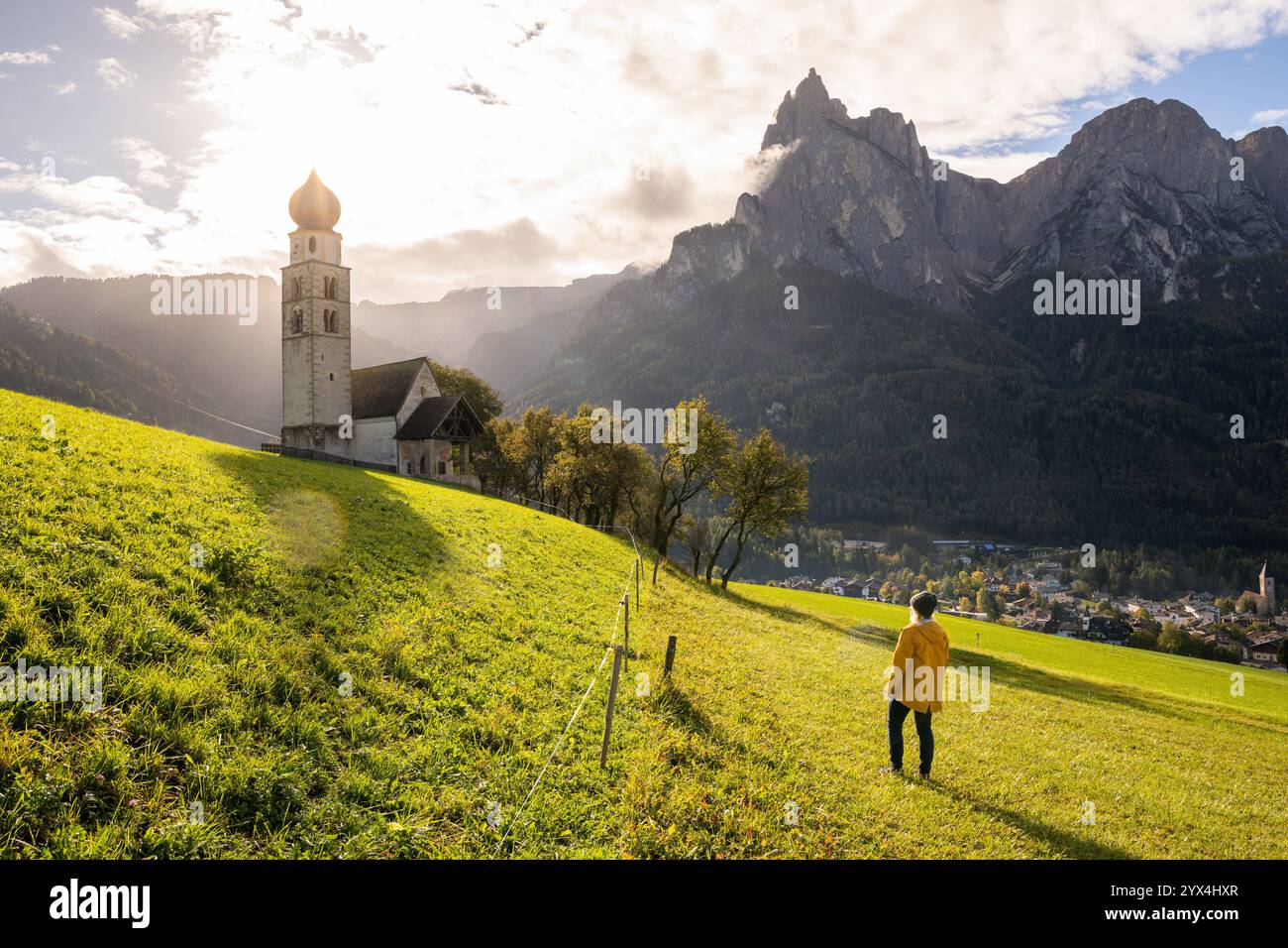 Frau in gelbem Regenmantel genießt Sonnenuntergang über den Dolomiten und der alten Steinkirche in Südtirol, Italien. Stockfoto