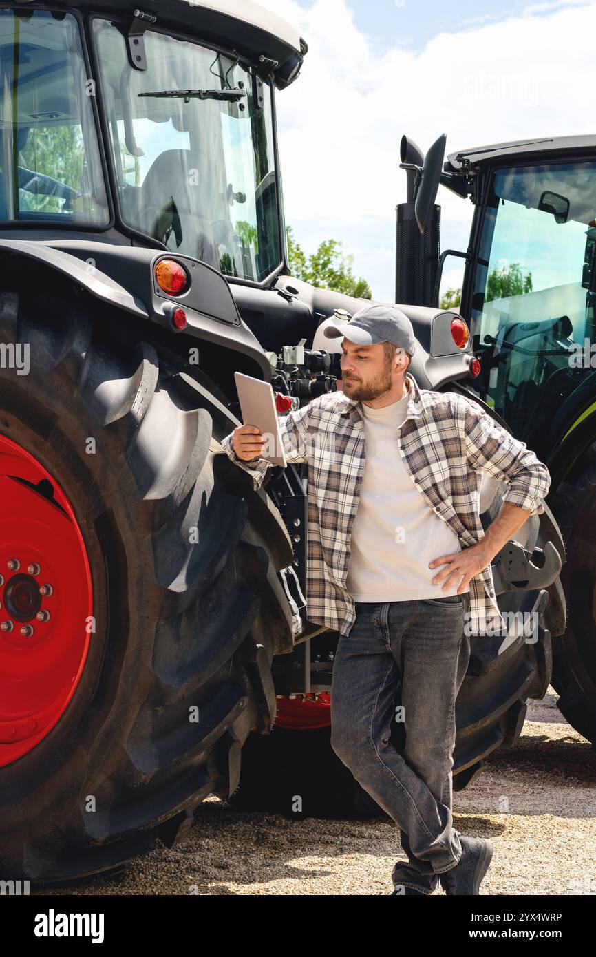 Kaukasischer Mann im karierten Hemd steht neben Traktoren und benutzt ein digitales Tablet. Stockfoto