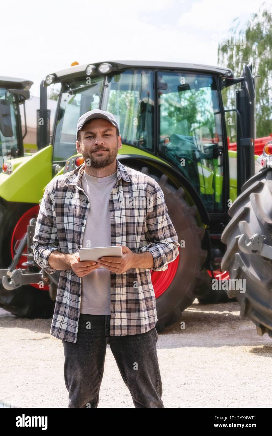 Kaukasischer Mann, der ein digitales Tablet auf dem Hintergrund von Traktoren im Landmaschinenhändler hält. Stockfoto