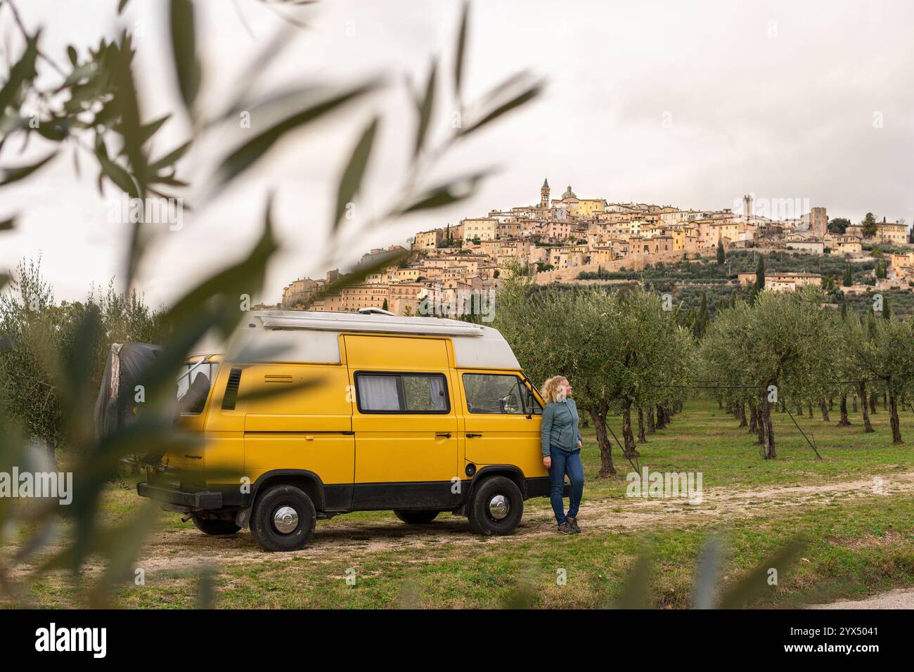 Junge Frau, die Umbrien in einem alten Wohnmobil erkundet, während sie durch Olivenhaine mit einer Bergstadt im Hintergrund fährt, Italien. Stockfoto