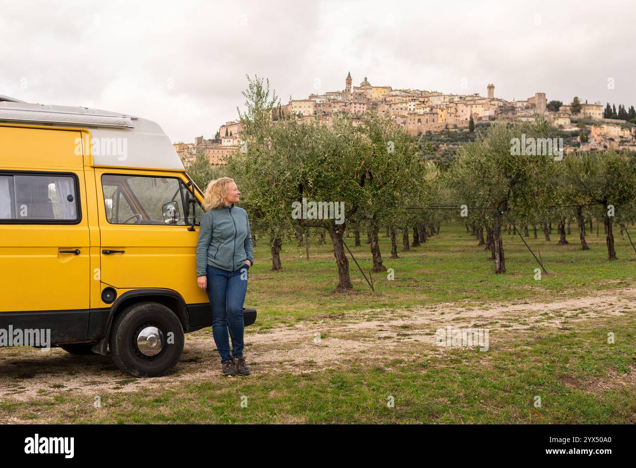 Junge Frau, die Umbrien in einem alten Wohnmobil erkundet, während sie durch Olivenhaine mit einer Bergstadt im Hintergrund fährt, Italien. Stockfoto