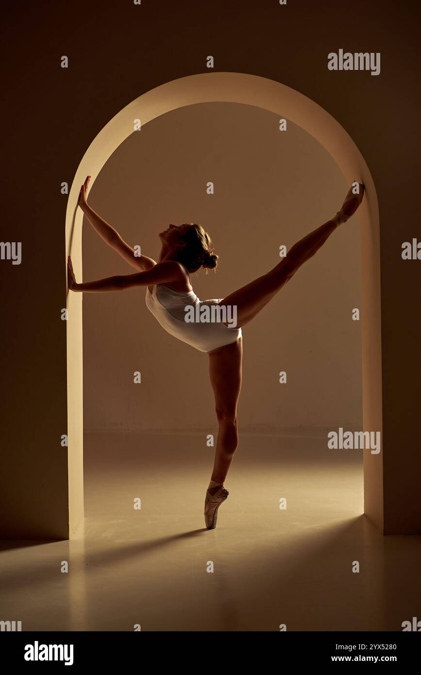 Ballerina, die sich in einem Bogengang erstreckt und in warmen Tönen beleuchtet ist. Konzept: Stärke, Flexibilität und zeitlose Ballettelegantheit. Stockfoto