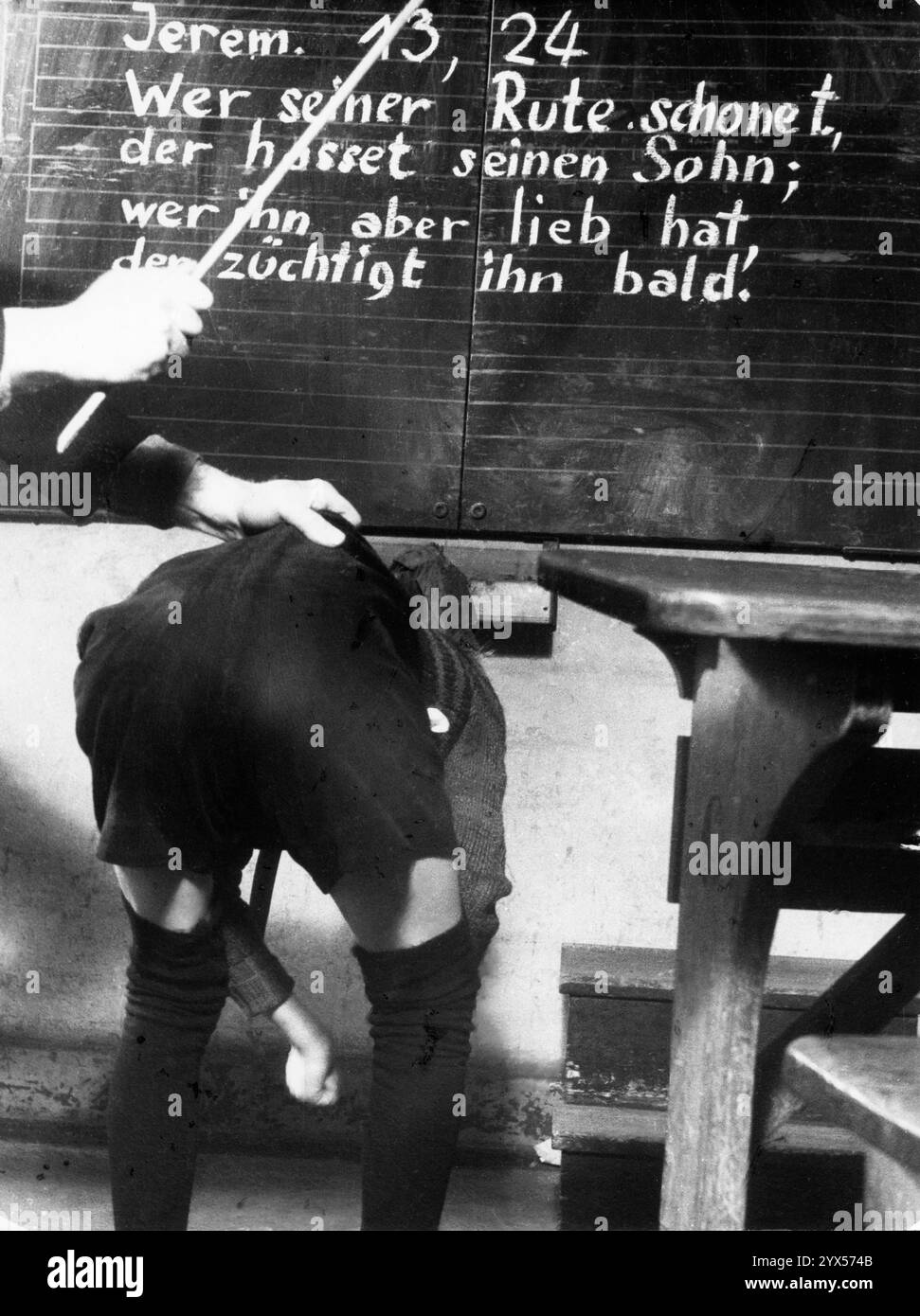 Ein Lehrer schlägt einem Schüler mit einem Stock auf den Rücken. Auf der Tafel steht ein Zitat aus der Bibel, Jer. 13:24: "Wer seinen Stab erspart, hasst seinen Sohn; wer ihn aber liebt, züchtigt ihn bald." [Automatisierte Übersetzung]“ Stockfoto