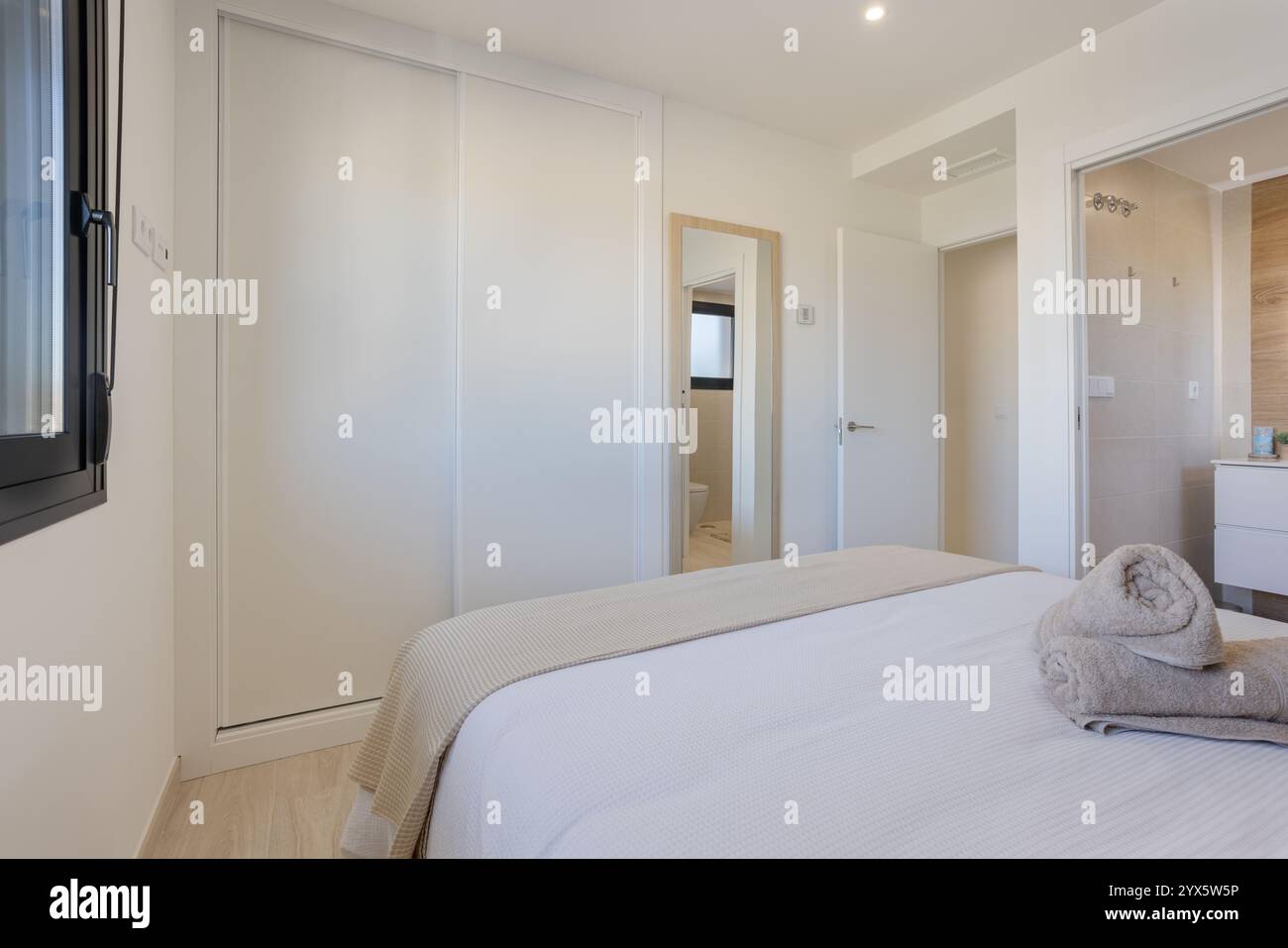 Modernes Schlafzimmer mit eingebautem Kleiderschrank, eigenem Bad, großem Fenster für natürliches Licht und gemütlicher, minimalistischer Einrichtung. Stockfoto