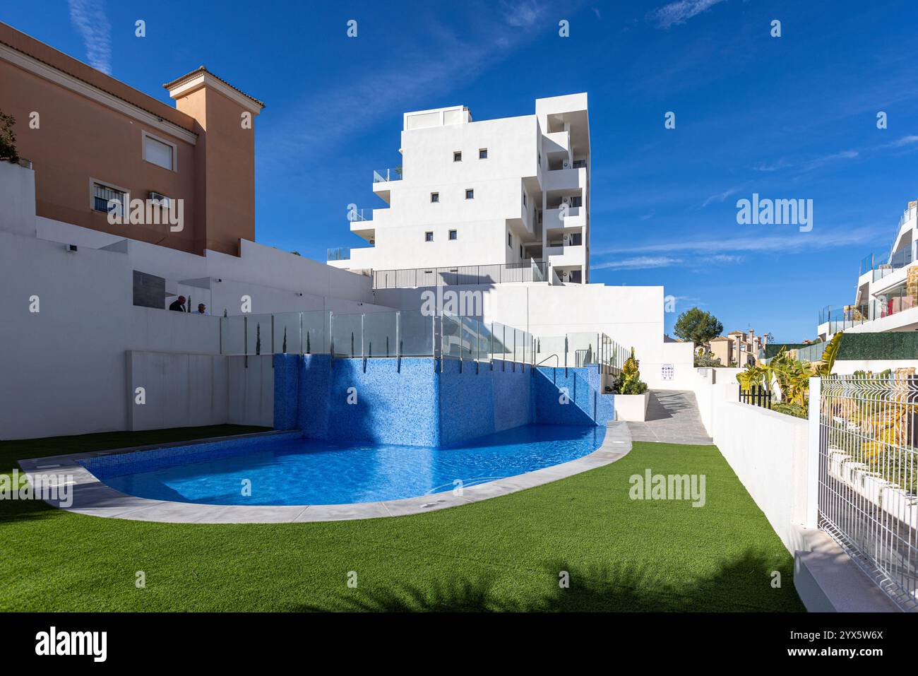 Ein stilvoller Wohnkomplex mit einem geschwungenen Pool mit blauen Fliesen, Kunstrasen und moderner Architektur unter sonnigem Himmel. Stockfoto