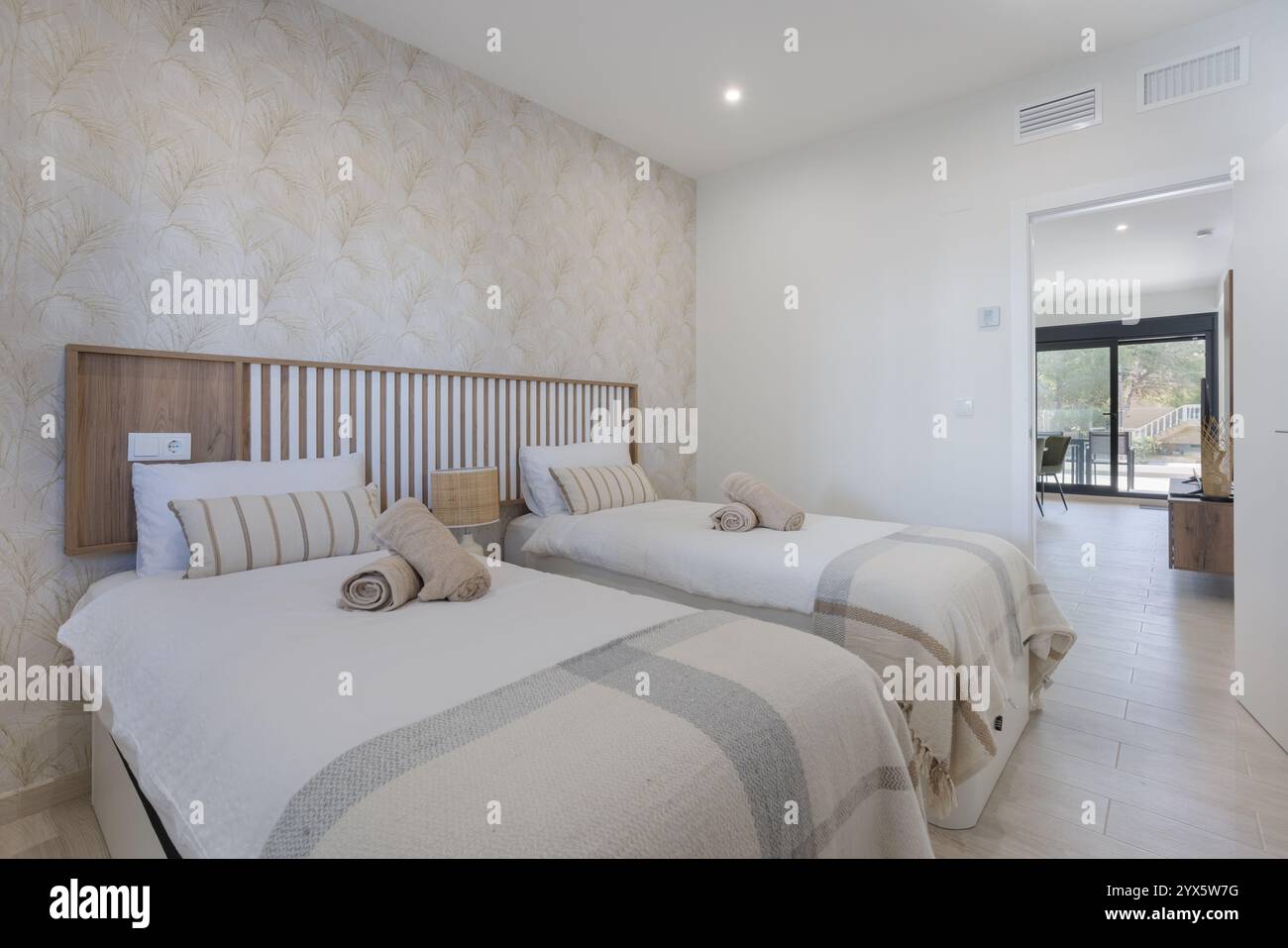 Elegantes Zweibettzimmer mit Holzkopfteilen, weichen neutralen Betten, gestreiften Kissen und natürlichem Licht, das von einer offenen Tür aus fließt. Stockfoto