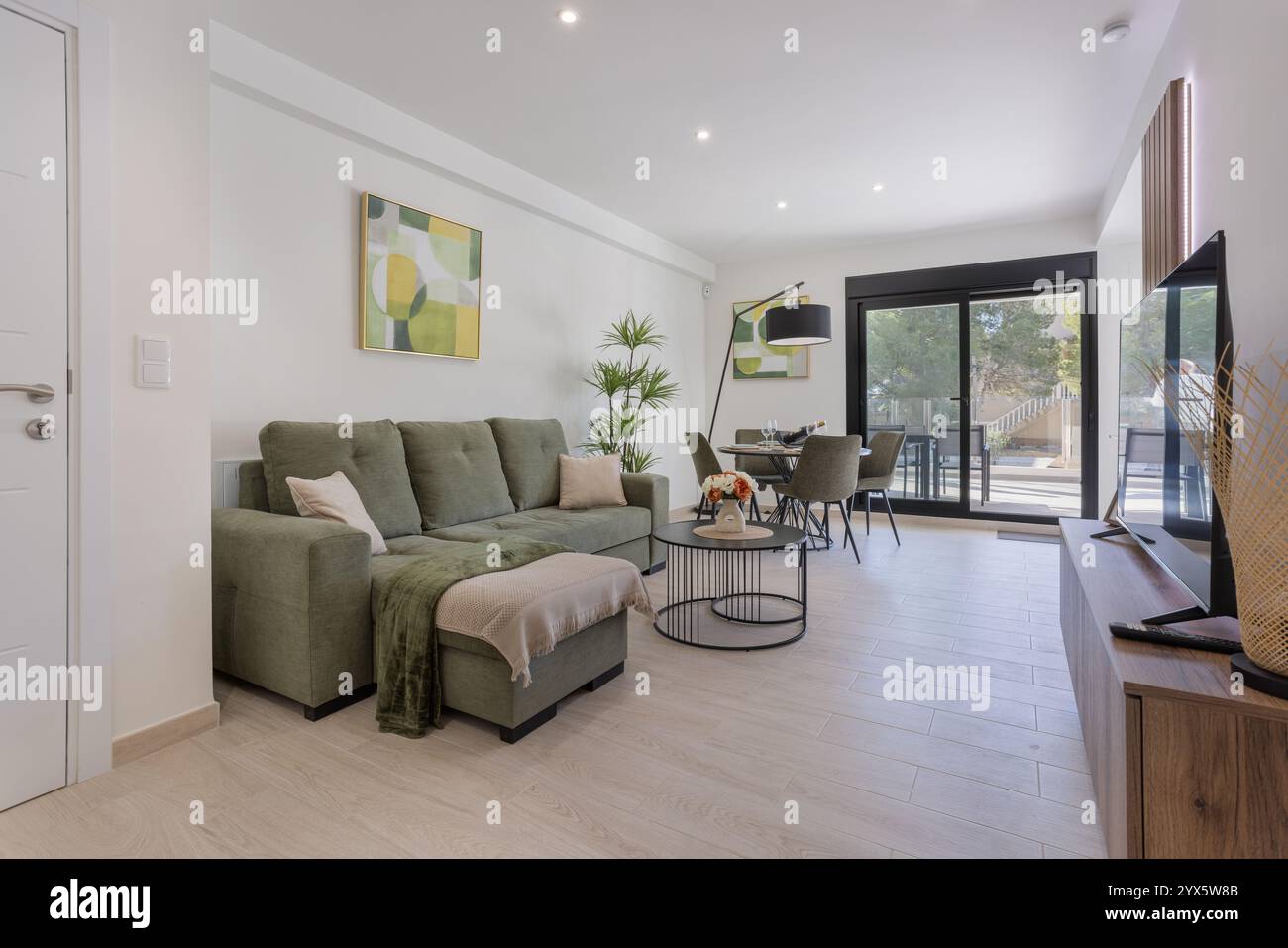 Helles und stilvolles, offen geschnittenes Wohn- und Esszimmer mit grünen Möbeln, minimalistischer Einrichtung und großen Schiebetüren, die auf eine Terrasse führen. Stockfoto