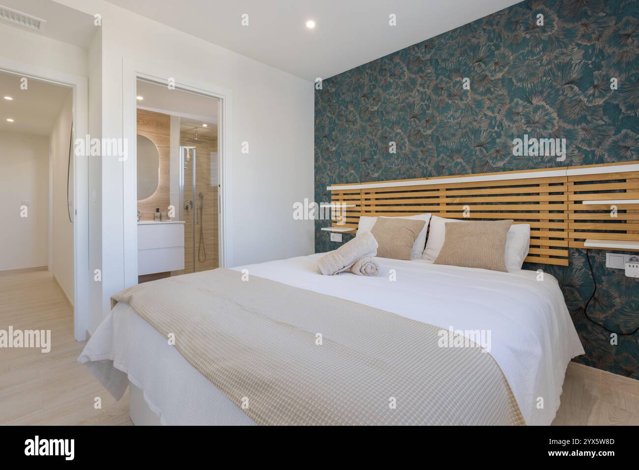 Modernes Schlafzimmer mit auffälligen geblümten Tapeten, Holzkopfteil, gemütlicher Bettwäsche und eigenem Bad mit minimalistischem Design. Stockfoto