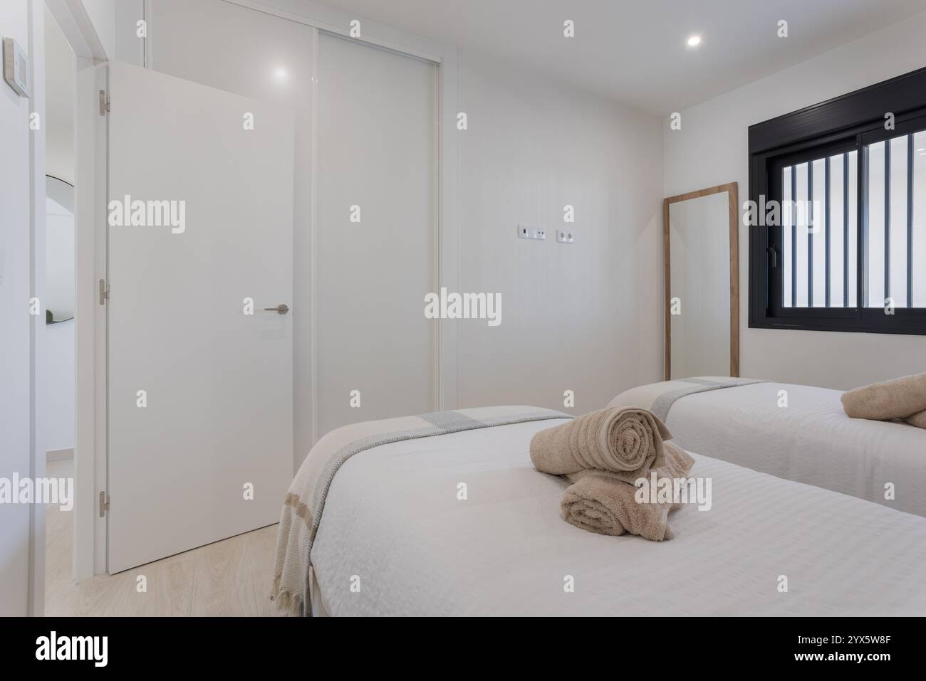 Minimalistisches Schlafzimmer mit Twin-Betten, frischer Bettwäsche, gerollten Handtüchern, großem Kleiderschrank und einem Fenster, das natürliches Tageslicht einlädt. Stockfoto