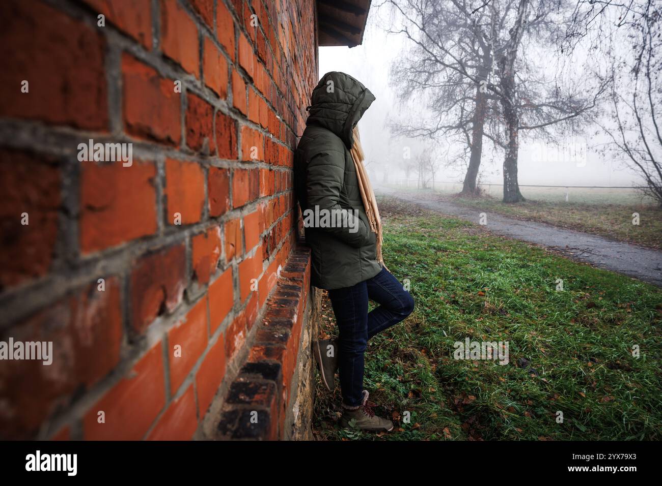 Traurige einsame Frau mit Kapuze, die im Nebel an einer Ziegelmauer steht. Einsamkeit und Depression in eigenen Gedanken Stockfoto