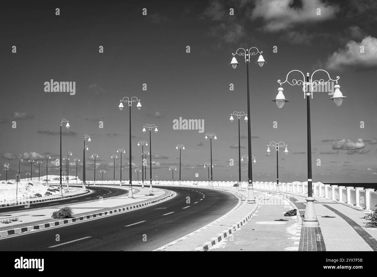 Ein fesselndes Schwarz-weiß-Bild von Mersa Matruhs ruhiger Küstenpromenade mit eleganten Straßenlaternen und einer geschwungenen Straße gegen den Himmel. Stockfoto