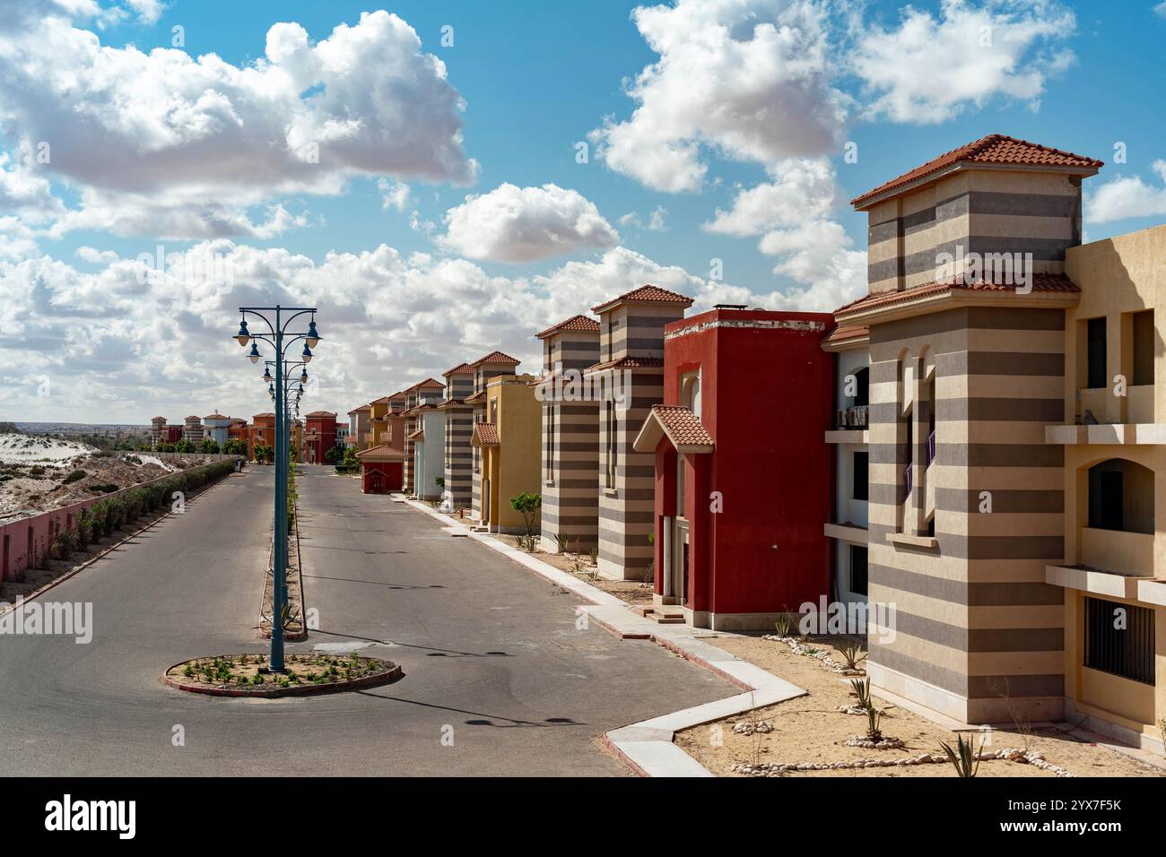 Farbenfrohe Villen mit gestreiften Fassaden und Ziegeldächern säumen eine ruhige Straße im Blue Porto Matruh Resort in Mersa Matruh, Ägypten, unter strahlendem blauen Himmel. Stockfoto