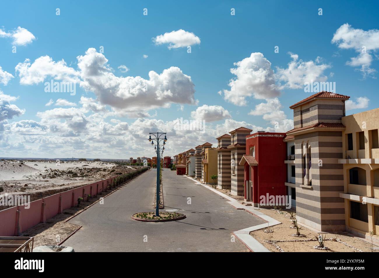 Farbenfrohe Villen mit gestreiften Fassaden und Ziegeldächern säumen eine ruhige Straße im Blue Porto Matruh Resort in Mersa Matruh, Ägypten, unter strahlendem blauen Himmel. Stockfoto