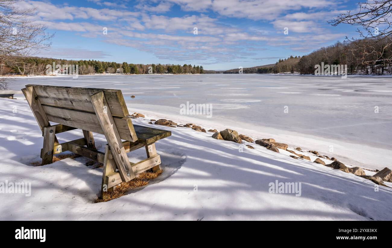 Eine leicht verwitterte Holzbank auf einem schneebedeckten Ufer liegt neben einem gefrorenen See in der Landschaft von Maryland. Stockfoto