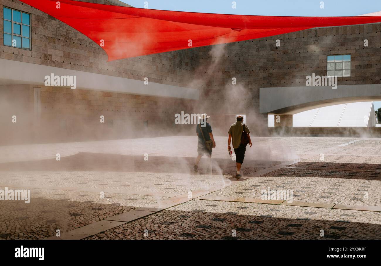 Zwei Personen genießen einen erfrischenden Nebel, während sie unter einem leuchtenden roten Schatten durch den Innenhof des Centro Cultural de belem in lissabon, portugal, spazieren Stockfoto