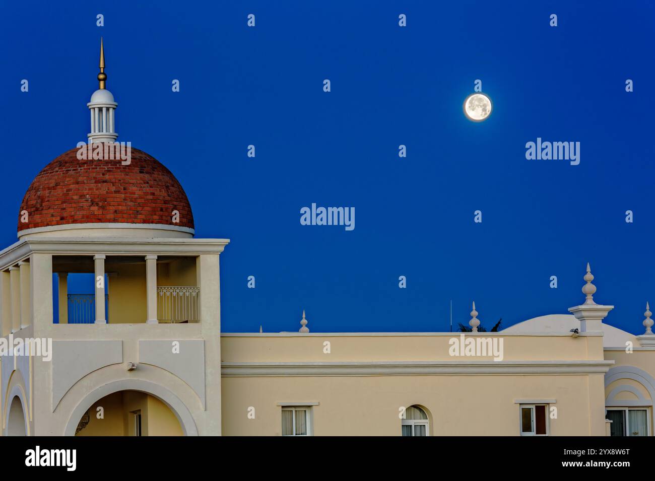 Ein großes weißes Gebäude mit einer roten Kuppel und einem großen Mond am Himmel. Das Gebäude ist von einem blauen Himmel umgeben Stockfoto