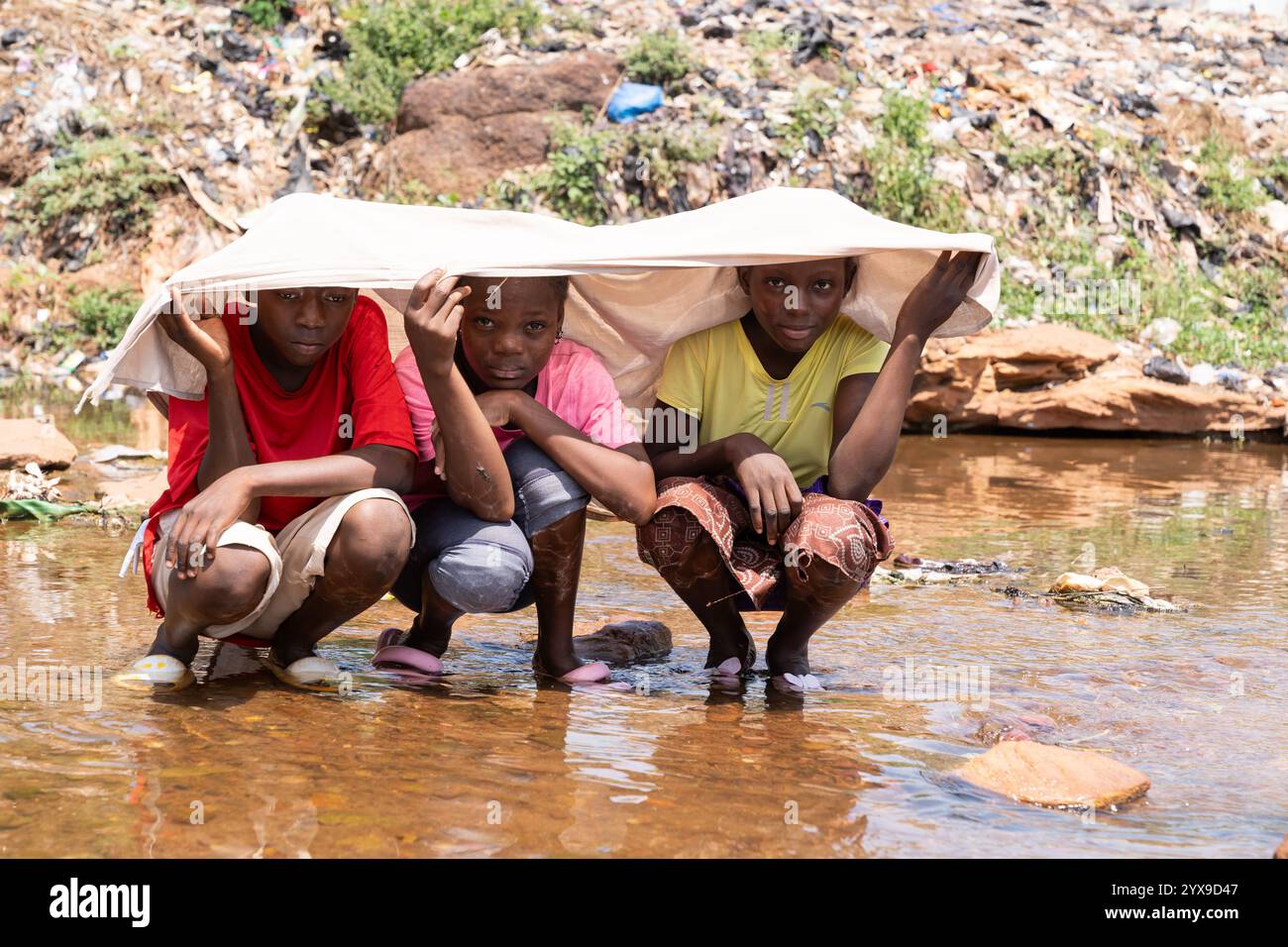 Gruppe afrikanischer Kinder, die sich vor der hellen Sonne unter einem Laken schützen und ihre Füße im Wasser kühlen. Klimawandel und globale Erwärmung Stockfoto