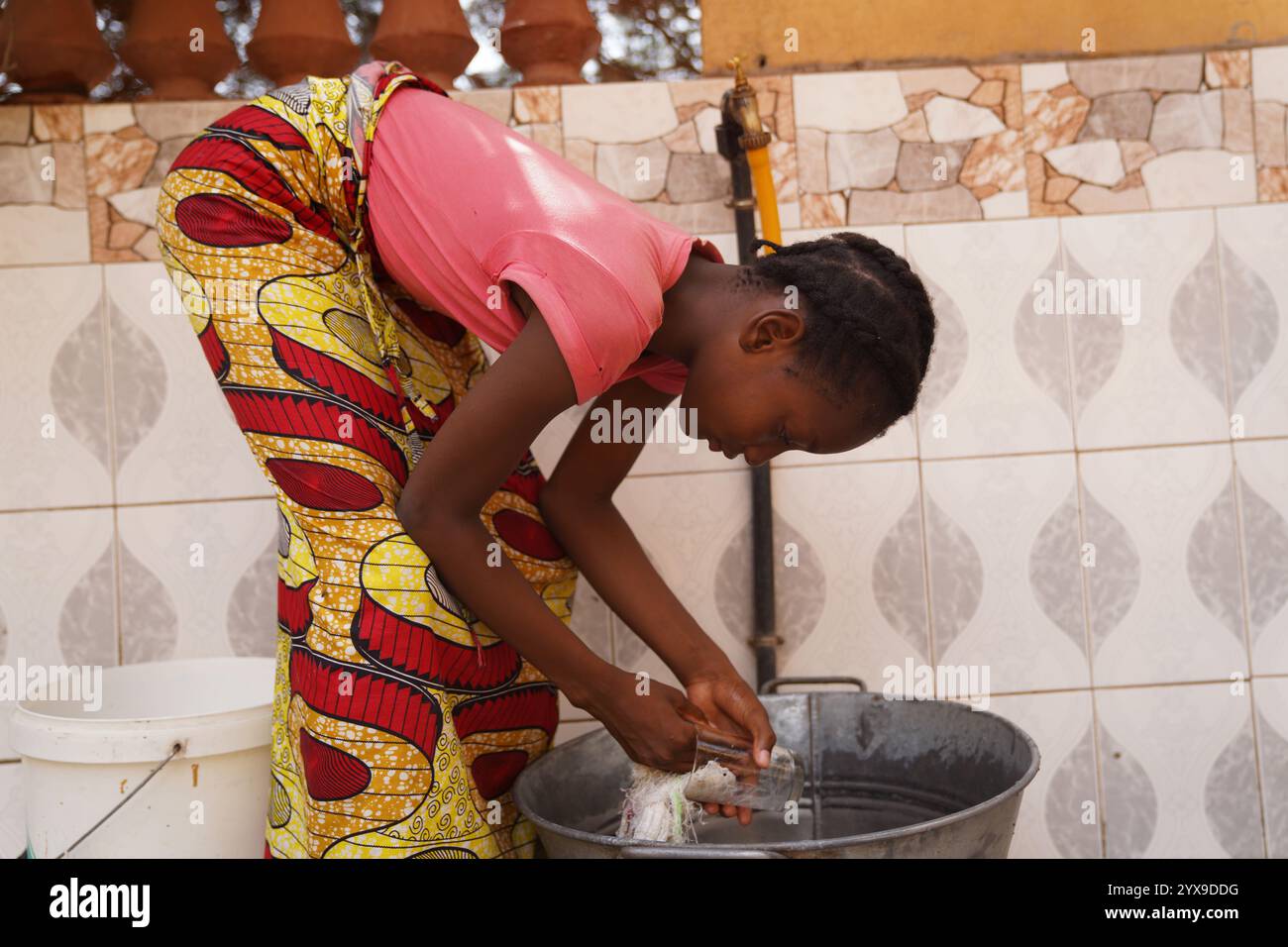 Junge afrikanische Mädchen in bunten Kleidern, die das Geschirr sorgfältig an einem Wasserhahn waschen Stockfoto