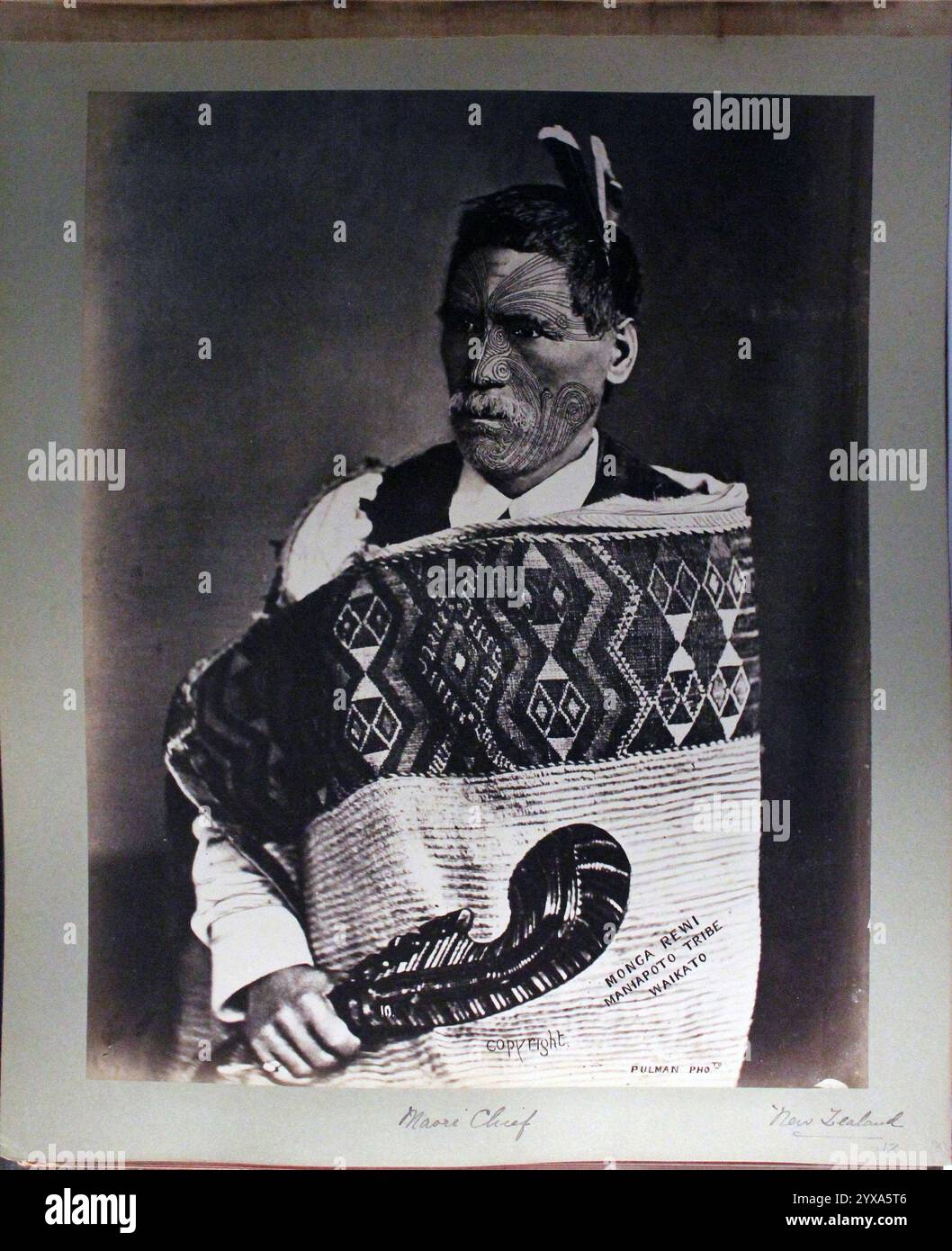 Porträt des Maori-Häuptlings Monga Rewi Maniapoto aus einer Publikation aus dem Jahr 1942 mit der Sammlung Victor Millard. Fotografiert von William Henry Thomas Partington um 1900, wahrscheinlich in Wanganui. Rewi posiert vor einem schlichten Hintergrund. Er hat einen dünnen weißen Bart, kurze, weiße Haare, die mit Federn verziert sind, und ausgedehnte Gesichtstattoos. Er trägt einen steifen Mantel mit geometrischen Mustern um seine Schultern, darunter ein europäisches Hemd und eine Weste. Er hält ein kurzes, geschnitztes Paddel in den Händen, möglicherweise ein Symbol der Autorität. Stockfoto