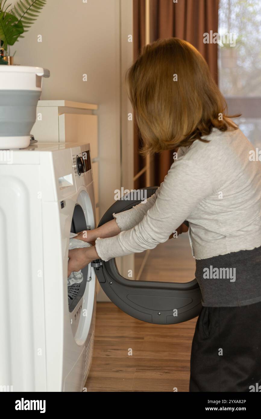 Eine Frau ist mit Waschaufgaben beschäftigt, indem sie Kleidung in eine Waschmaschine lädt. Der Innenraum ist hell und modern, mit sanfter Beleuchtung und einem Hauch von G Stockfoto