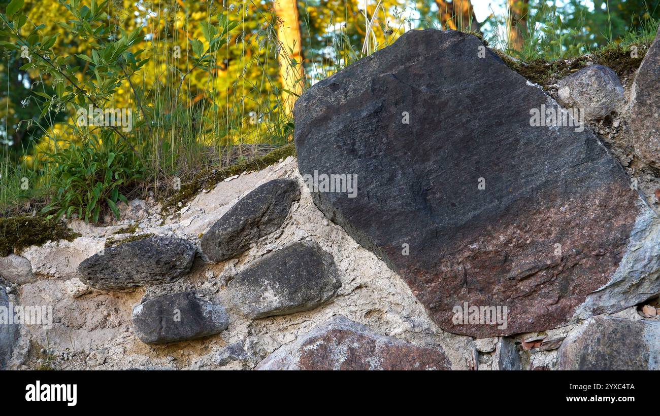 Nahaufnahme eines alten Steinmauerfragments, teilweise mit wildem Gras und Pflanzen bewachsen. Die Textur der Steine und die natürliche Überwucherung sind hoch Stockfoto