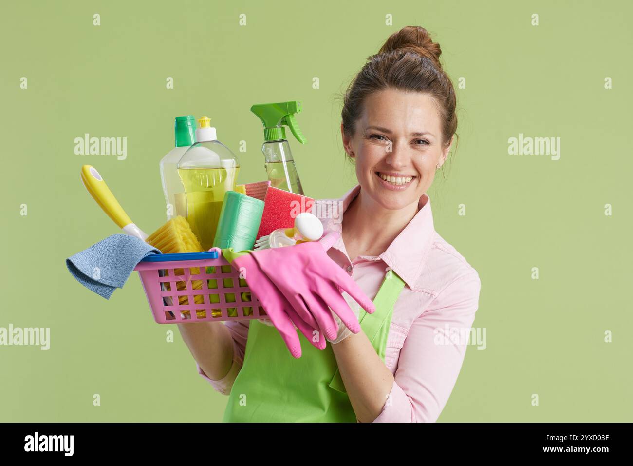 Frühjahrsputz. Glückliche moderne Hausfrau mittleren Alters in grüner Schürze und Gummihandschuhen isoliert auf Grün mit Korb mit Reinigungsprodukten. Stockfoto