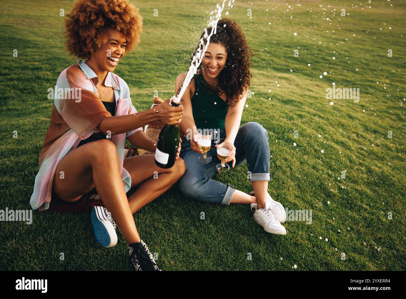 Zwei junge Freunde sprühen aufgeregt Champagner im Park und feiern mit Lachen und Freude an einem sonnigen Tag, umgeben von üppigem Gras und einem lebhaften Atmo Stockfoto
