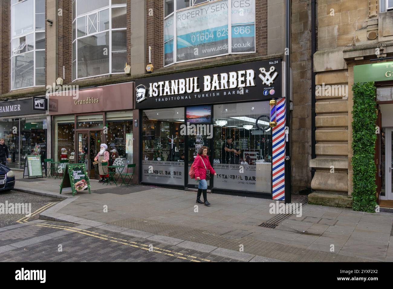 Frau in rotem Mantel, die an Instanbul Barber, einem traditionellen türkischen Friseurladen, in St. Giles St, Northampton, Großbritannien, vorbeiläuft Stockfoto