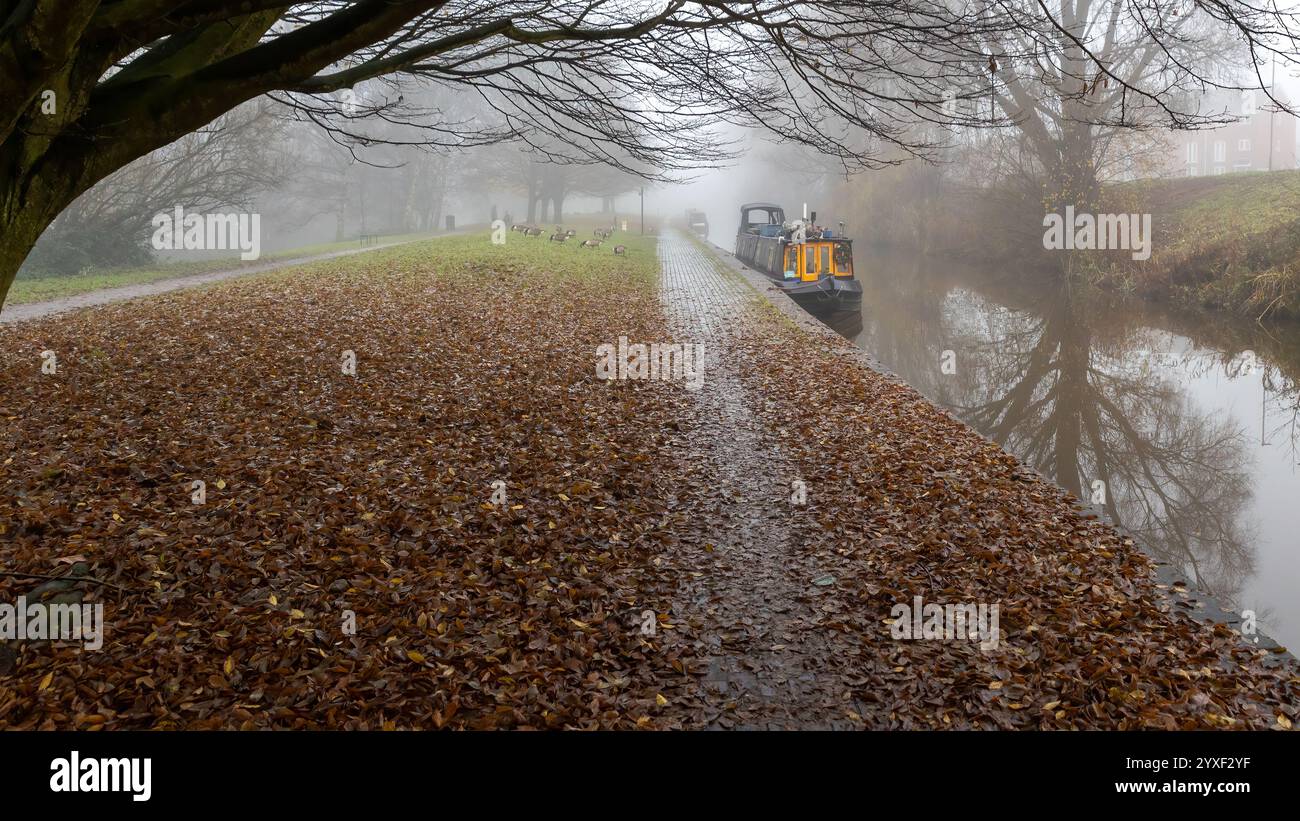 Aufgenommen im Herbst mit Blick auf einen Kanalschleppweg. Die braunen Blätter bedecken den Boden. In der Ferne liegen enge Kanalboote, die im leichten Nebel vertäut sind. Stockfoto