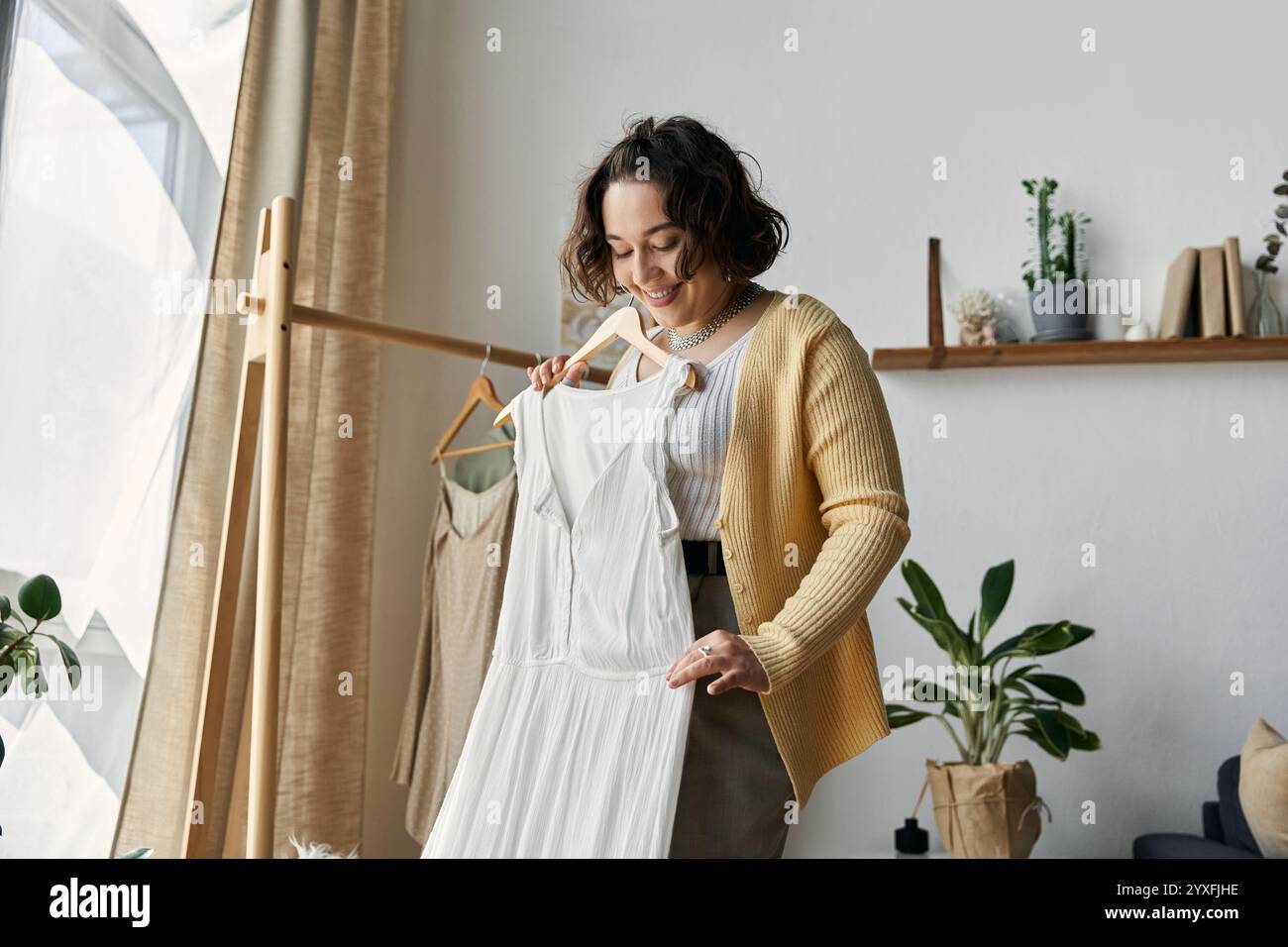 Eine junge Frau mit lockigen Haaren schaut sich in ihrem lebendigen Wohnraum gerne ein Kleid an. Stockfoto