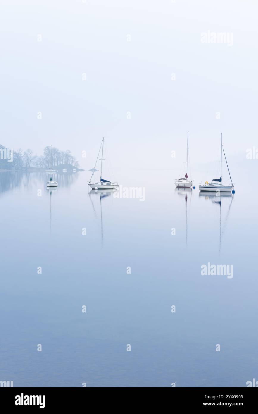 Ruhiger See mit spiegelartigen Reflexen und Yachten an einem nebeligen Morgen. Windermere, Lake District, Großbritannien. Stockfoto