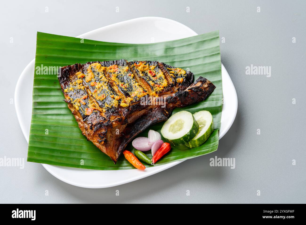 Ikan Pari Bakar oder gegrillter Sting Ray in Chili-Currysauce serviert auf Bananenblatt mit Schalotten, Gurke und Chili. Stockfoto