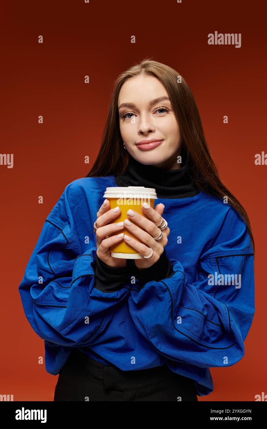 Eine lebendige Frau in einem blauen Pullover trinkt Kaffee vor einem warmen orangen Hintergrund. Stockfoto