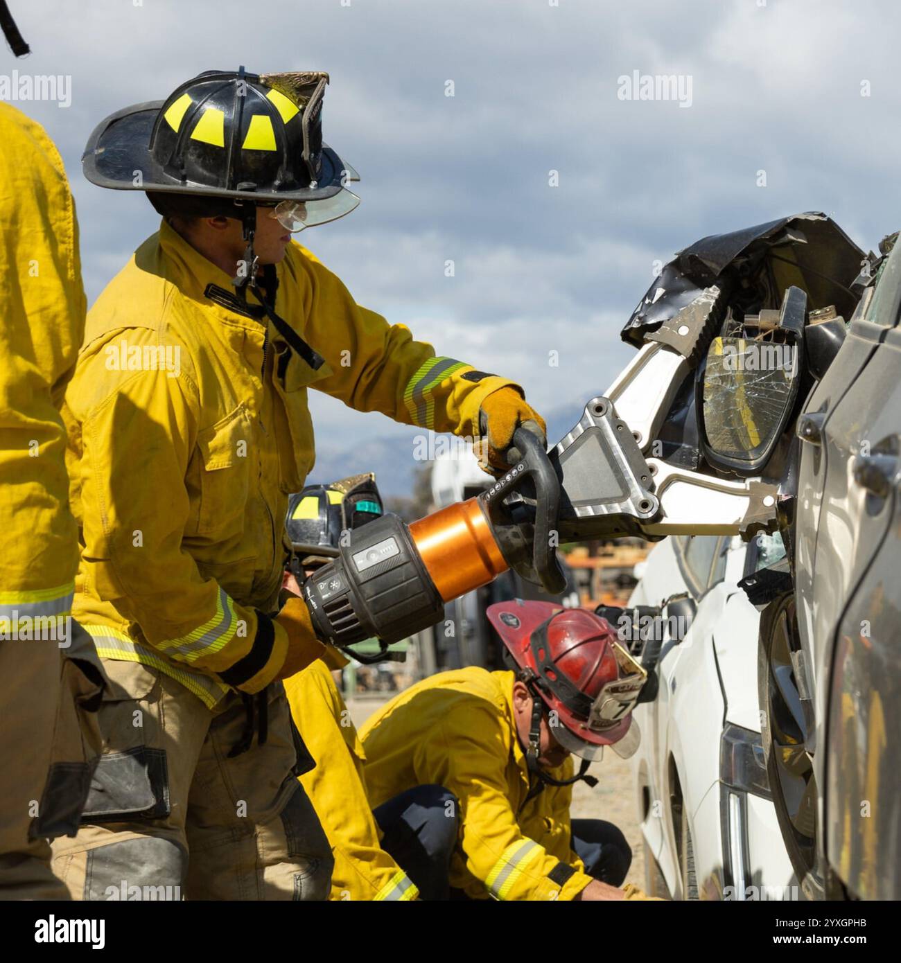 Feuerwehrleute in Aktion mit hydraulischen Werkzeugen, um eine Fahrzeugrextraktion durchzuführen, bei der Teamarbeit und Rettungsaktionen unter bewölktem Himmel gezeigt werden Stockfoto