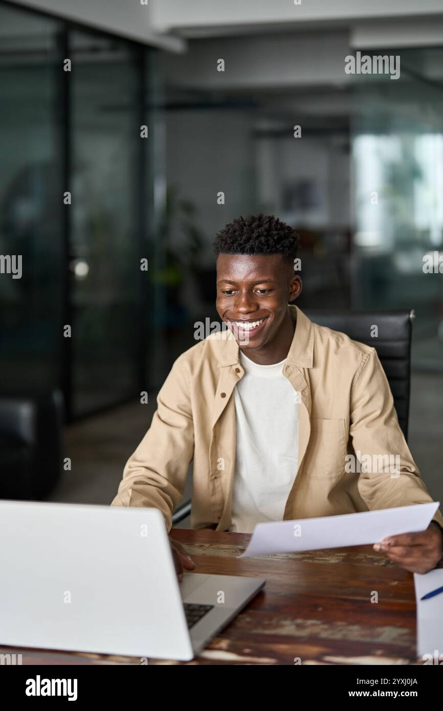 Glücklicher junger afrikanischer Geschäftsmann, der Dokumente mit Laptop auf der Arbeit hält. Stockfoto
