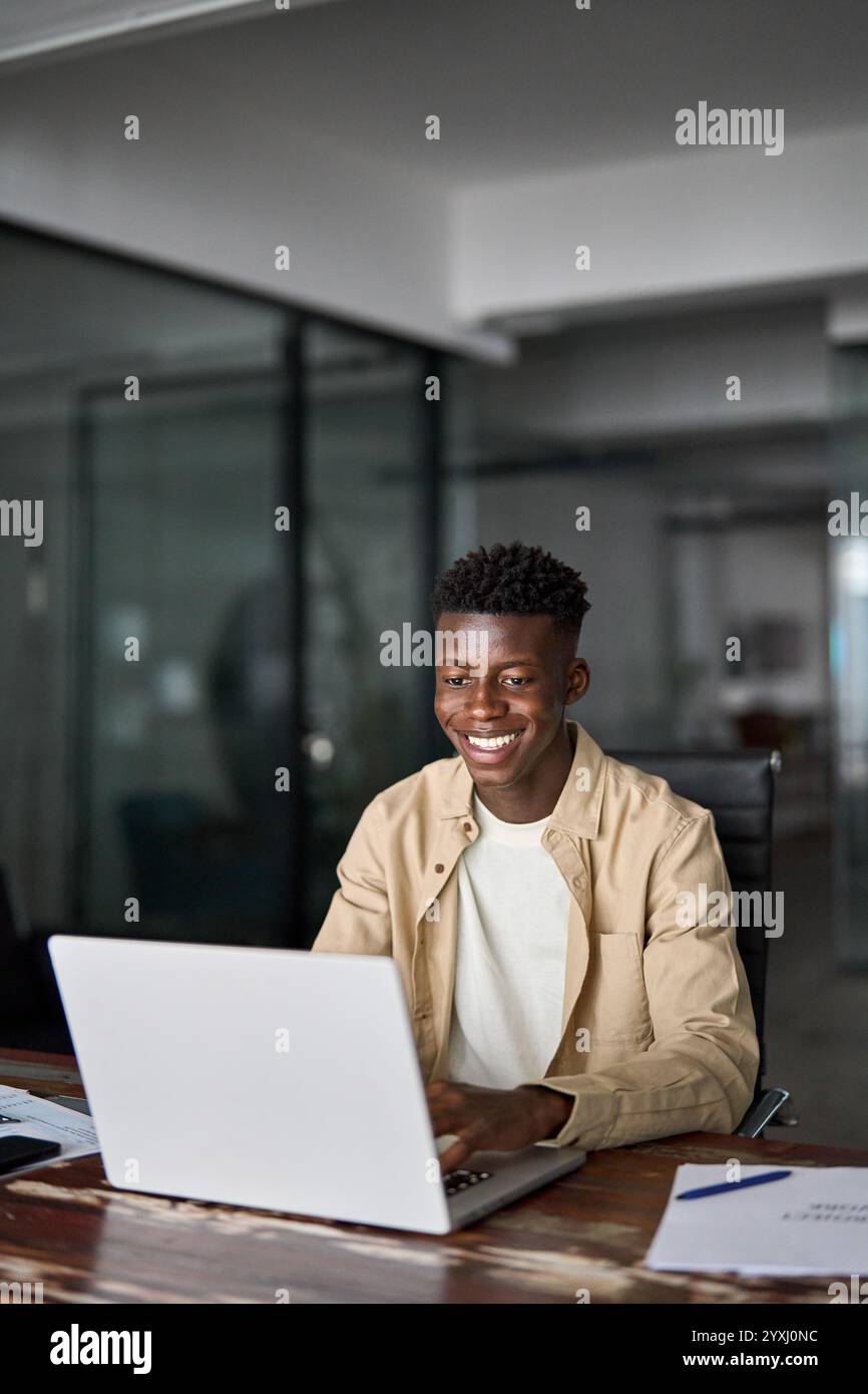 Hektischer afrikanischer Geschäftsmann, der im Büro mit einem Laptop arbeitet. Vertikales Foto. Stockfoto