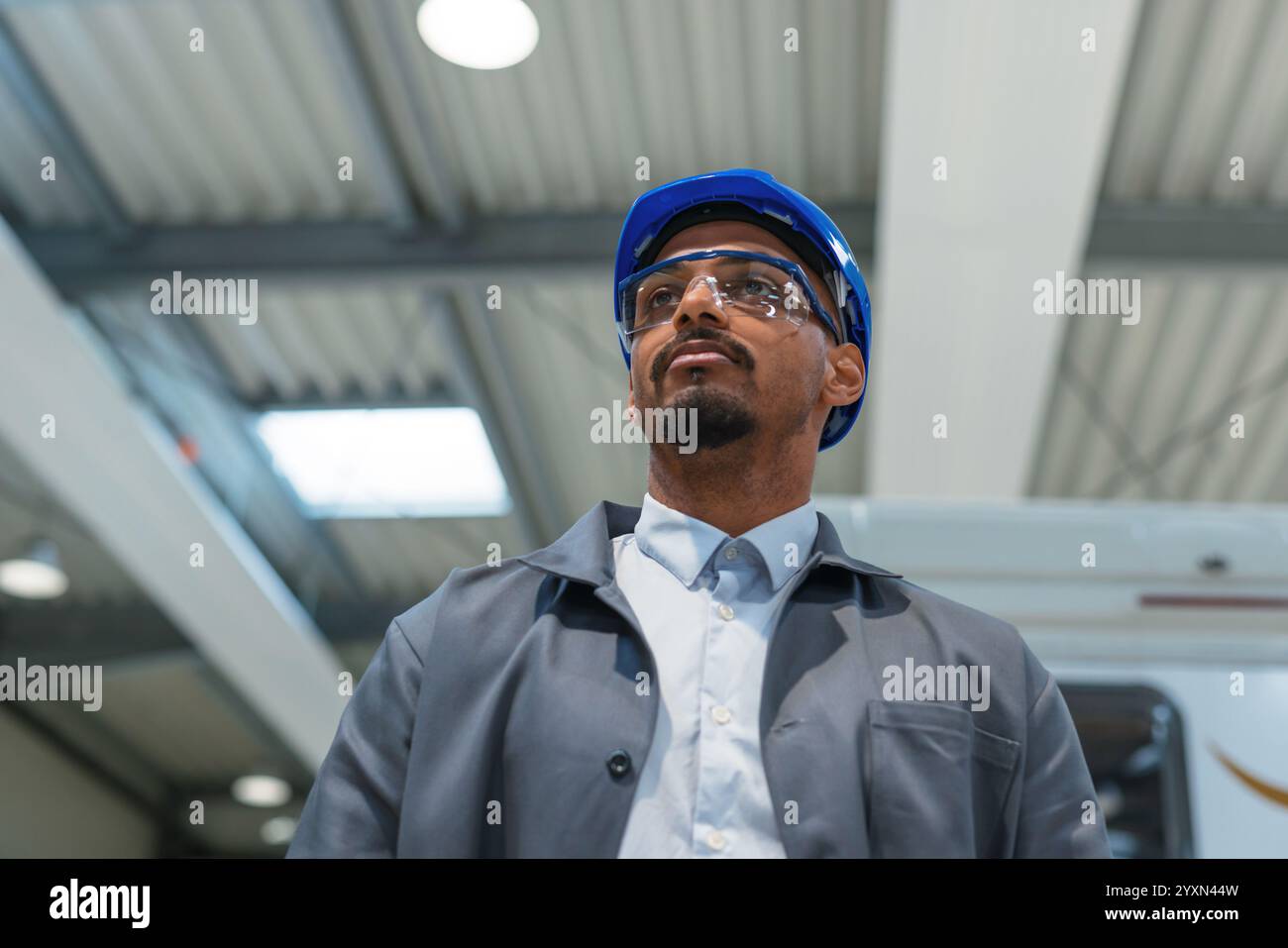 Afroamerikanischer Ingenieur, der einen grauen Arbeitsmantel und einen blauen Helm trägt und die Produktion und Programmierung einer Maschine in einer Fabrik steuert. Stockfoto