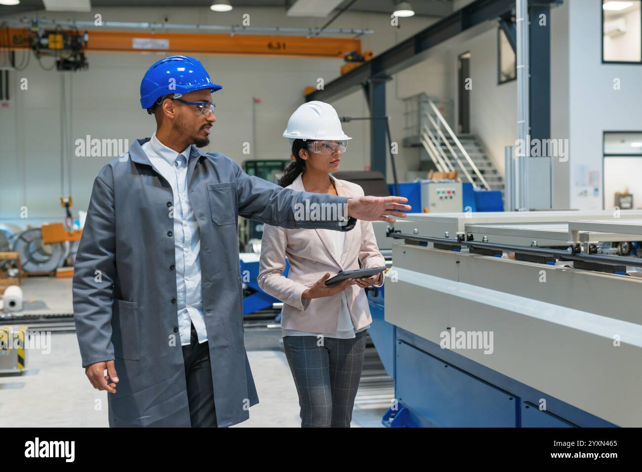 Afroamerikaner Ingenieur und eine Inderin, eine Fabrikleiterin, die ein Tablet hält, durch die Produktionslinie läuft, Inspektionen durchführt, Vorderansicht. Stockfoto