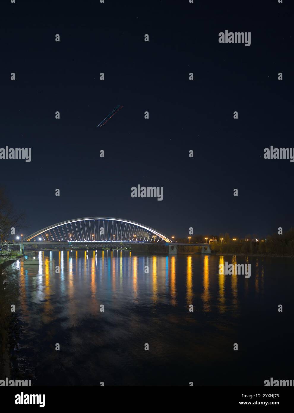 Eine hell beleuchtete moderne Brücke bei Nacht mit farbenfrohen Reflexen auf dem Fluss vor einem klaren dunklen Himmel. Stockfoto