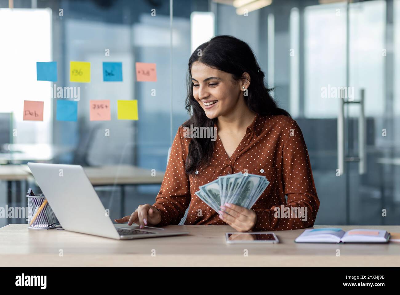 Eine junge Frau, die an einem Schreibtisch sitzt und mit einem Laptop Geld hält und lächelt. Erfasst Themen wie Erfolg, Unternehmergeist, Online-Arbeit, Finanzen und Geschäft in einem modernen, dynamischen Umfeld am Arbeitsplatz. Stockfoto