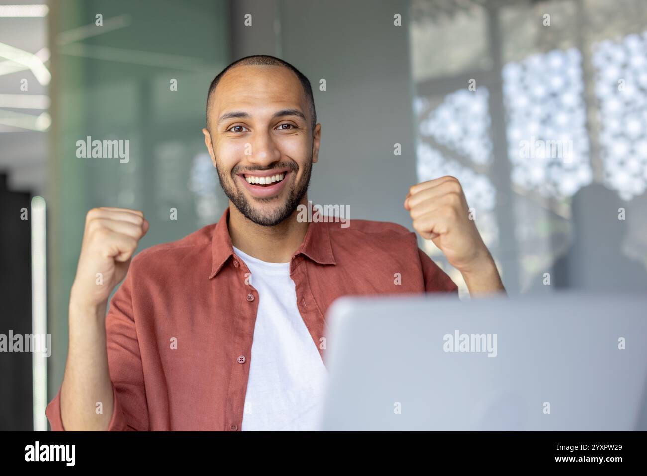 Eine fröhliche Person in einer Büroumgebung, die Freude mit gestreckten Fäusten ausdrückt und Erfolg und Positivität repräsentiert. Der lichtdurchflutete Hintergrund ergänzt das übertragene Leistungsgefühl. Stockfoto