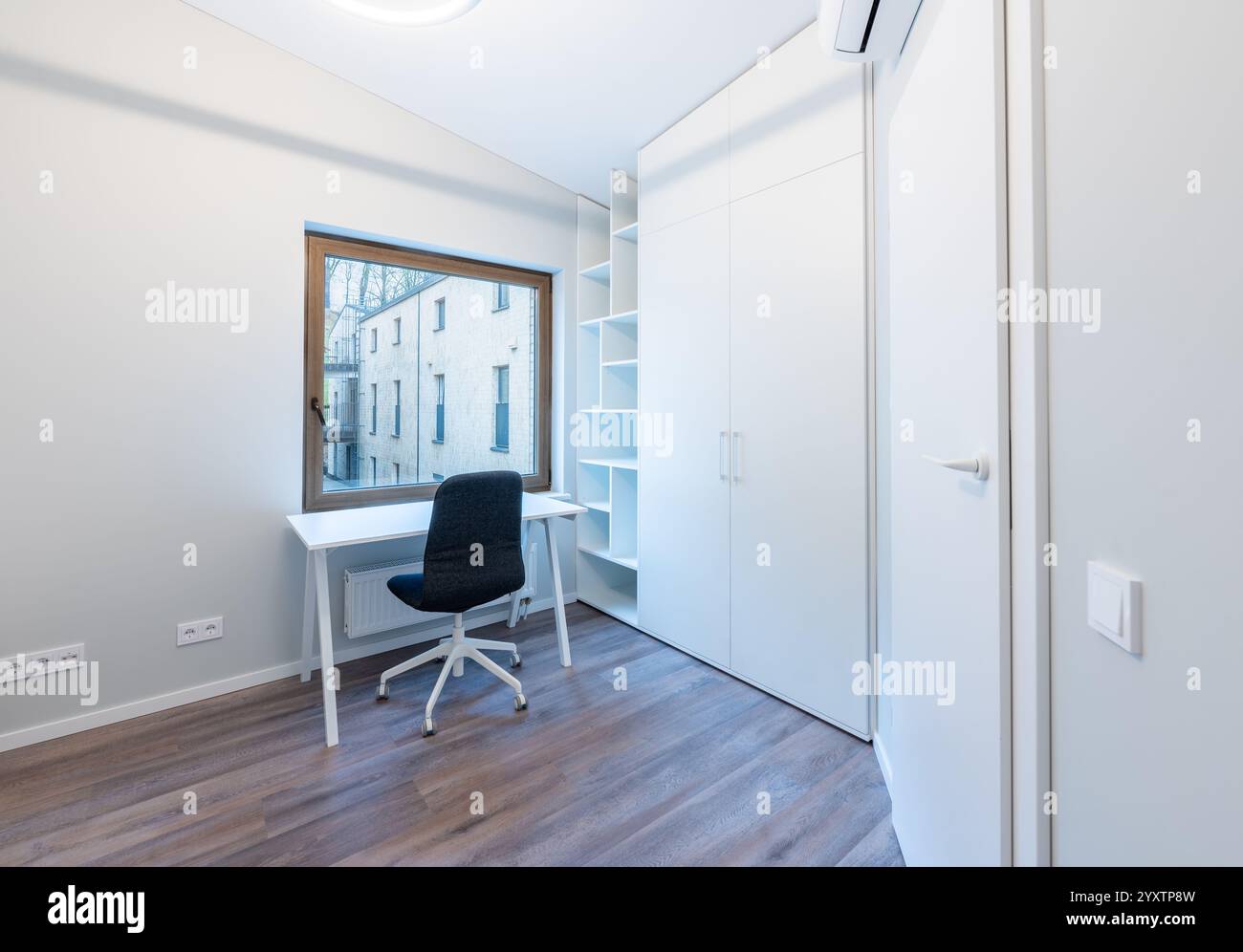 Helles und geräumiges Home Office mit weißem Schreibtisch und Stuhl, großem Fenster mit Blick auf den Außenbereich und minimalistischen Stauräumen, Klimaanlage an der Wand. Modern Stockfoto