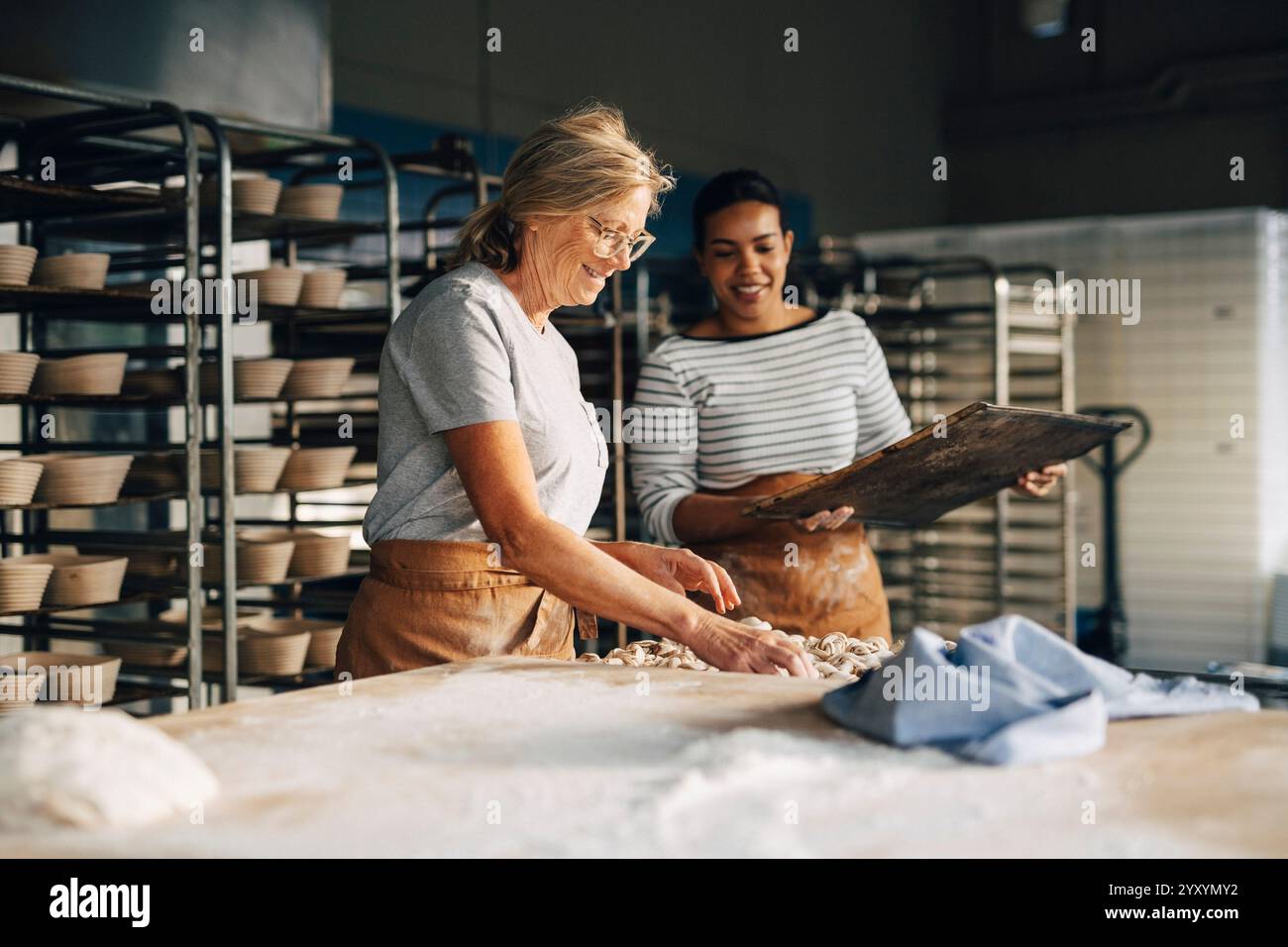 Lächelnde ältere Bäckerin arrangiert Zimtbrötchen von einer Kollegin, die ein Tablett hält, während sie in der Bäckerei arbeitet Stockfoto