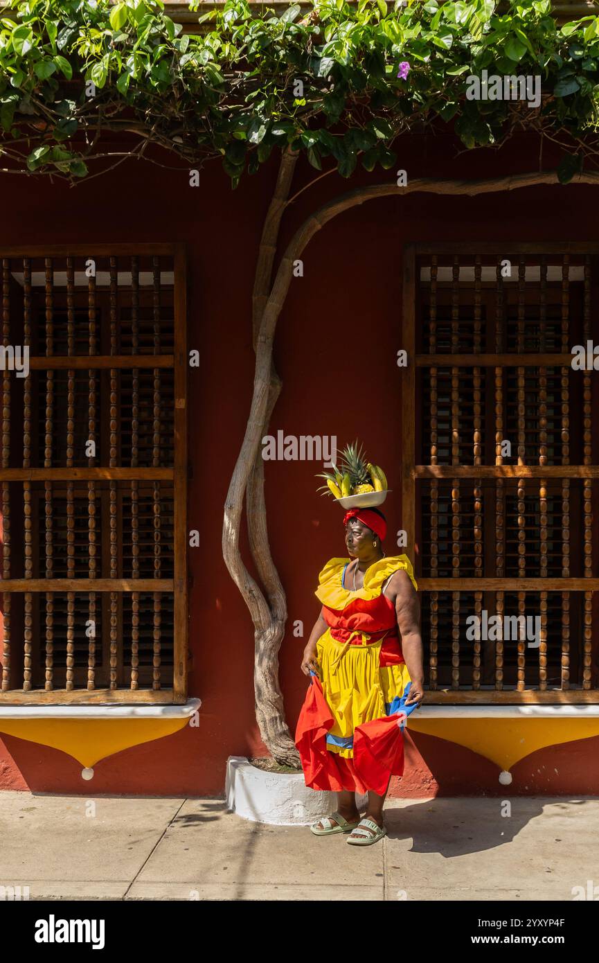 Eine Palenquera-Frau in Einem bunten Kleid mit Einem Obstkorb auf dem Kopf, die vor Einem Kolonialgebäude mit Holzfenstern steht Stockfoto