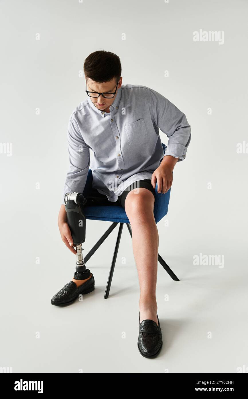 Ein junger Mann mit Brille sitzt auf einem blauen Stuhl und passt seine Beinprothese mit einem fokussierten Blick an. Stockfoto