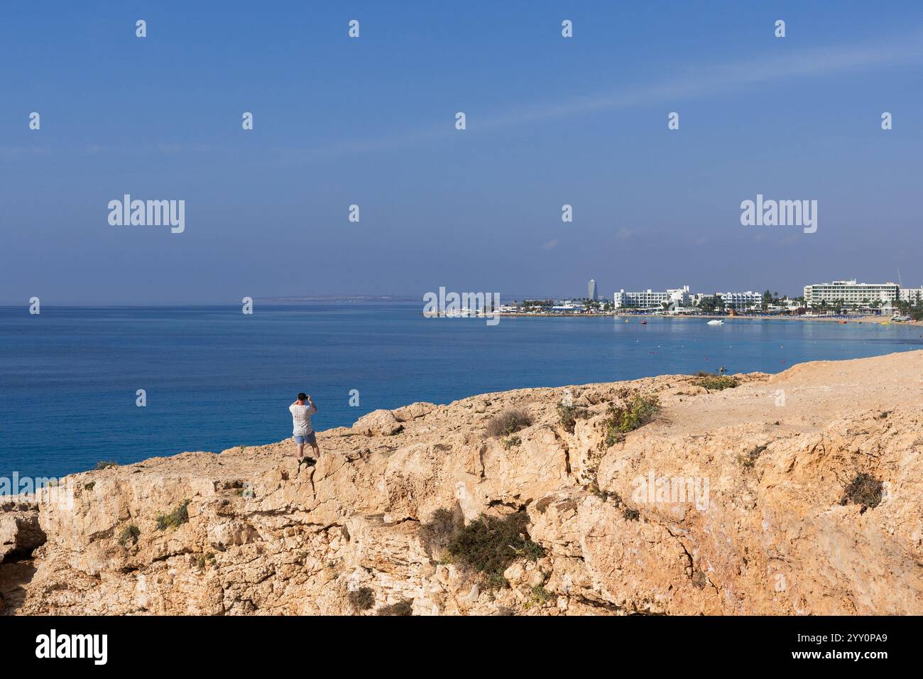 Kleine Figur eines Mannes, der mit dem Rücken auf einem riesigen dunklen Felsen vor dem Hintergrund des Meeres und des klaren blauen Himmels bleibt. Perfekte Aussicht. Reisekonzept Stockfoto