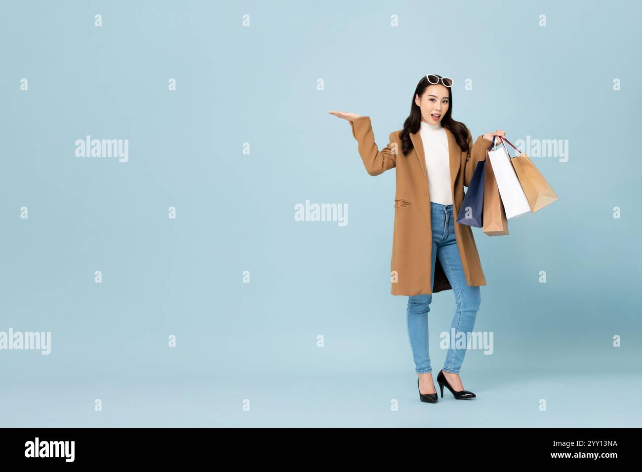 Schöne asiatische Frau, die Wintermantel trägt, Einkaufstaschen mit geöffneter Handgeste in hellblauer Farbe isolierten Studiohintergrund hält Stockfoto