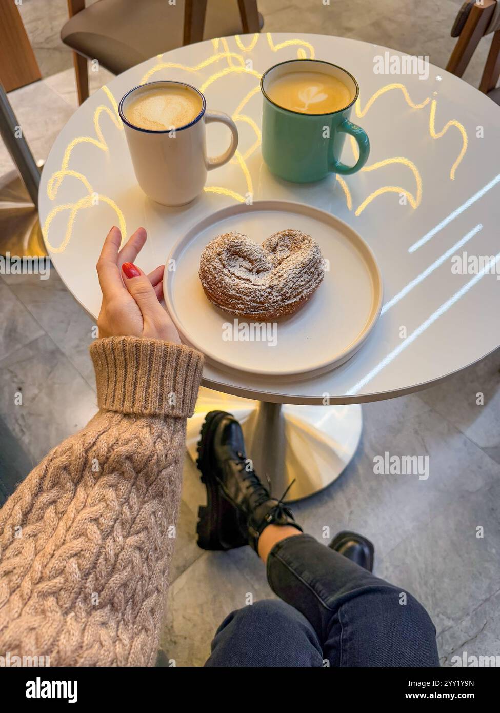 Dieses Foto fängt eine ruhige Winterszene in einem stilvollen Café ein. Eine kuschelige Hand in einem beigefarbenen Strickpullover reicht zu einem Tisch, der zwei warme Getränke hält o Stockfoto