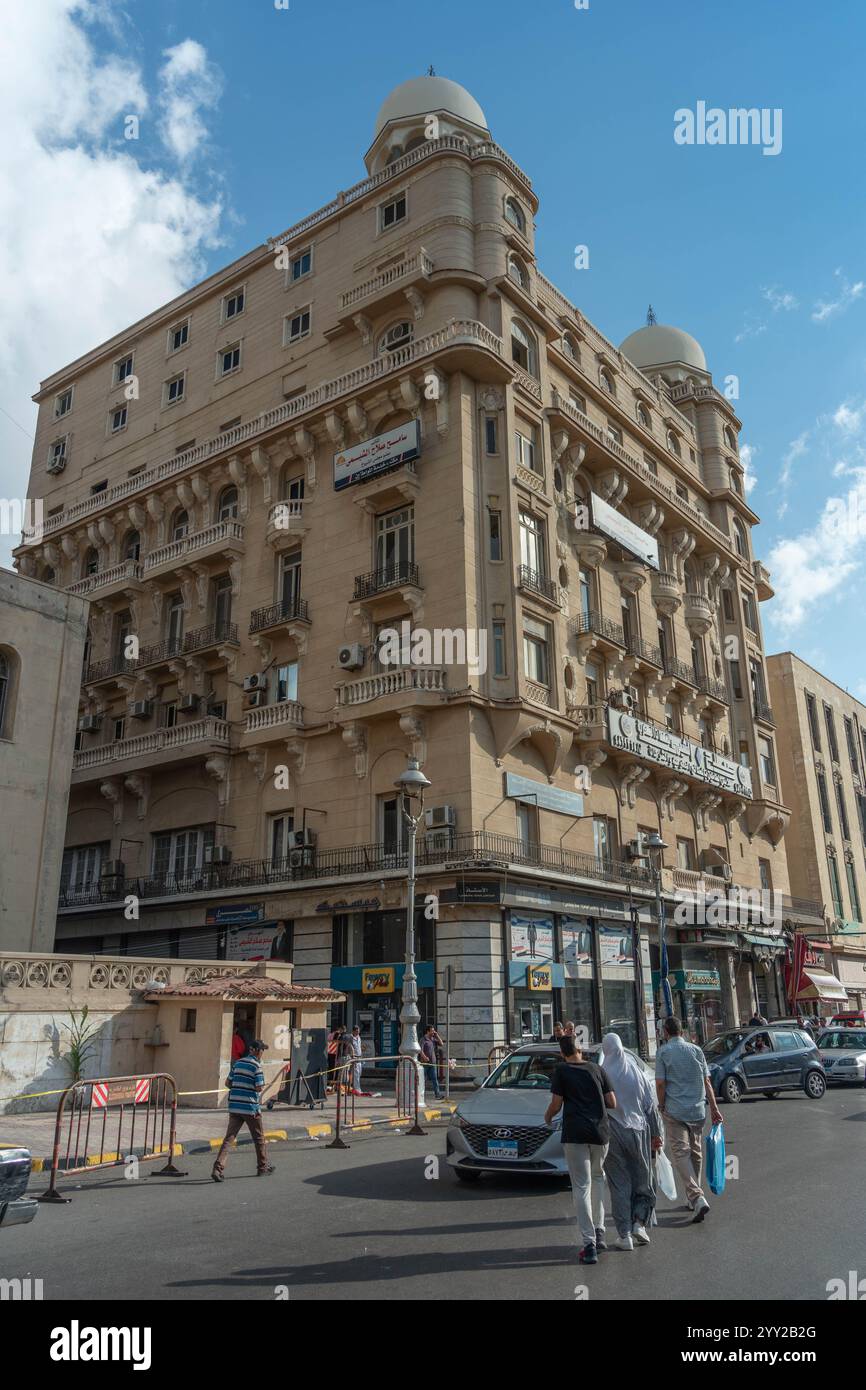 Historisches architektonisches Gebäude mit kunstvollen Balkonen, Kuppeldach und beigefarbener Fassade in Alexandria, Ägypten, mit Straßenaktivitäten und Fußgängern. Stockfoto